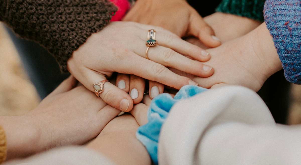 L'image montre plusieurs mains jointes, laissant apparaître des manches et des pulls colorés, suggérant un groupe de personnes réunies dans un esprit d'unité ou de soutien.