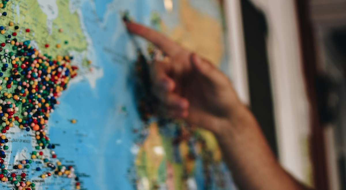 Une carte du monde colorée avec de nombreux petits points représentant des lieux ou des points d'intérêt, et une main pointant vers une zone précise sur la carte.