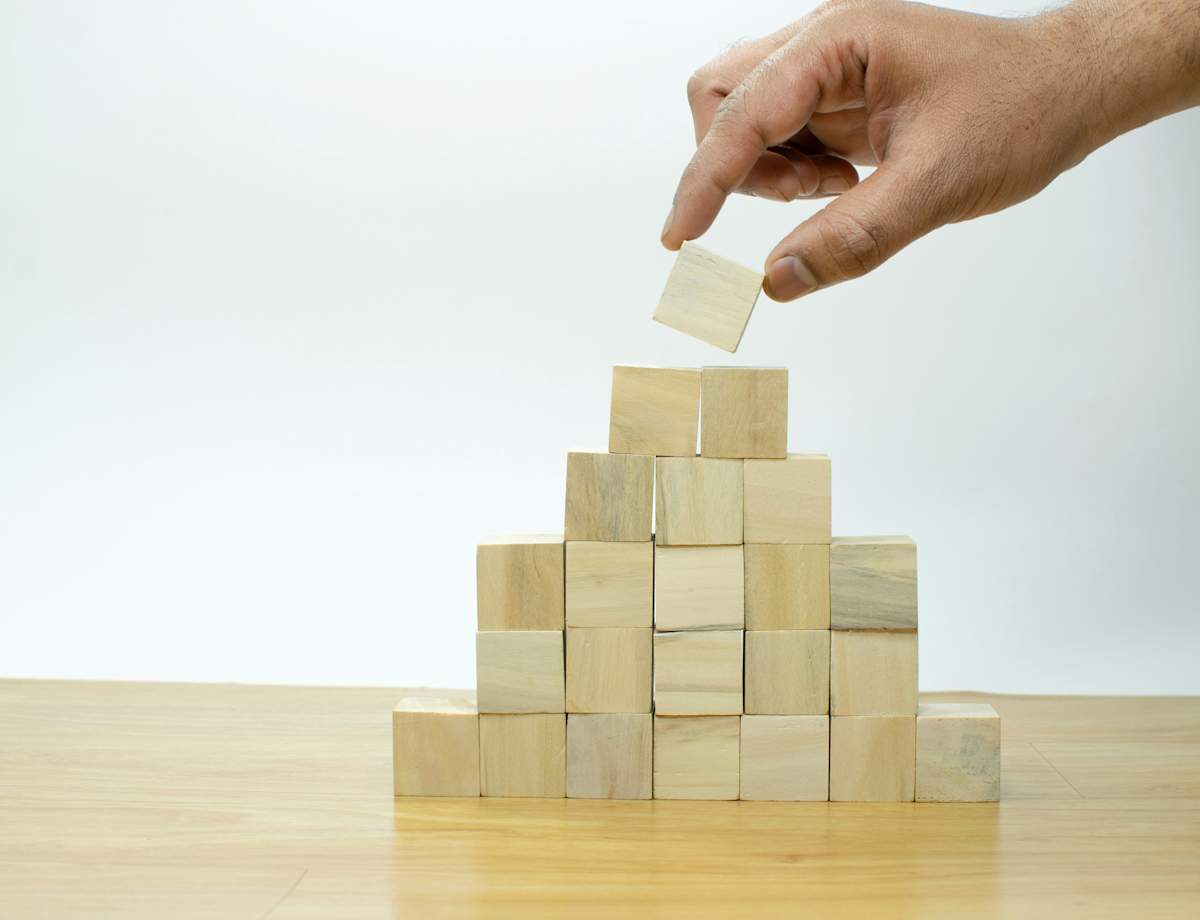 Une main dépose délicatement un bloc de bois au sommet d'une structure pyramidale composée d'autres blocs de bois, posée sur une table en bois.