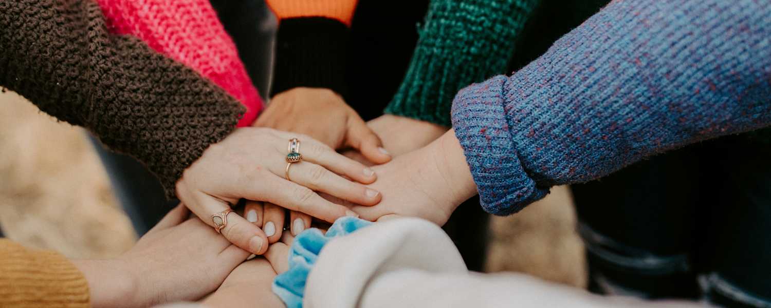 L'image montre plusieurs mains jointes, laissant apparaître des manches et des pulls colorés, suggérant un groupe de personnes réunies dans un esprit d'unité ou de soutien.