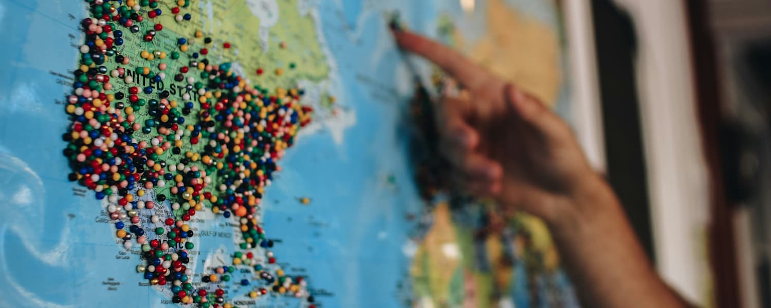 Une carte du monde colorée avec de nombreux petits points représentant des lieux ou des points d'intérêt, et une main pointant vers une zone précise sur la carte.