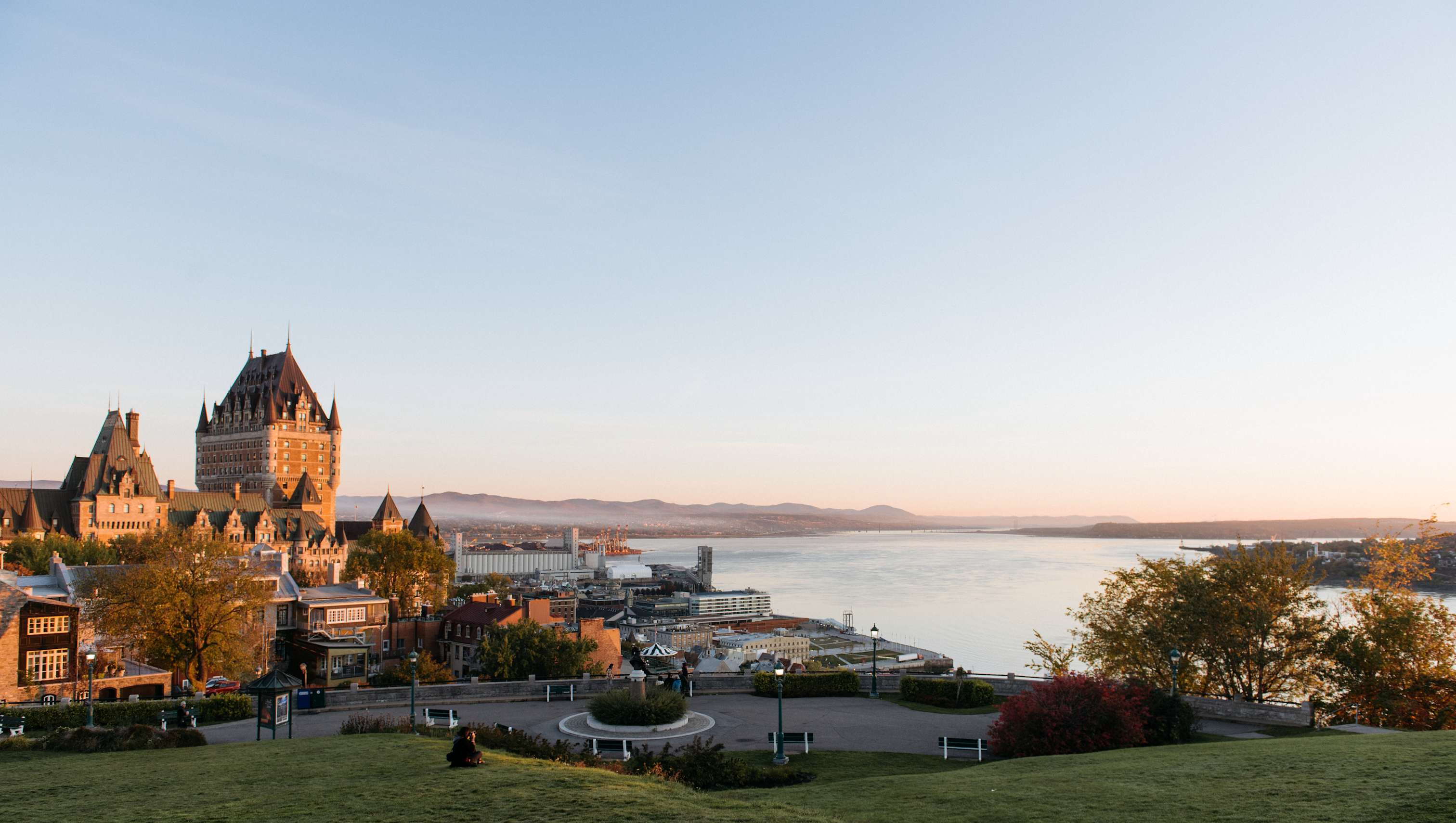 Un paysage urbain pittoresque avec une imposante structure ressemblant à un château surplombant une étendue d'eau, entourée d'une végétation luxuriante au premier plan.