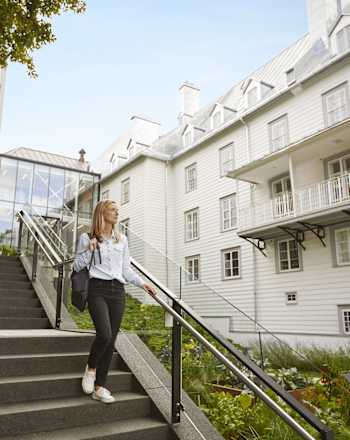 Une jeune femme en tenue décontractée monte un escalier menant à un immeuble résidentiel de plusieurs étages avec un balcon et entouré de verdure.