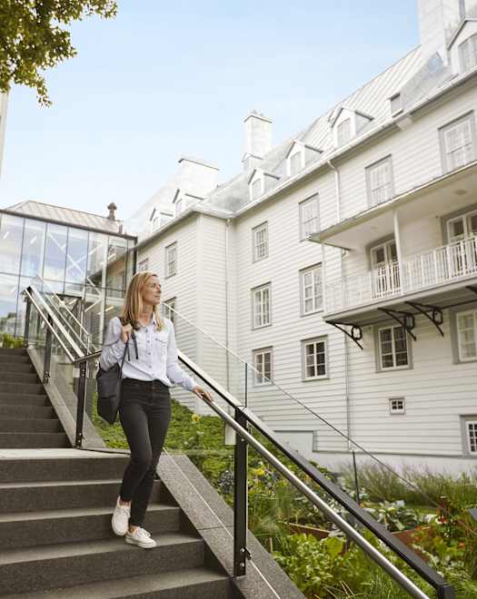 Une jeune femme en tenue décontractée monte un escalier menant à un immeuble résidentiel de plusieurs étages avec un balcon et entouré de verdure.