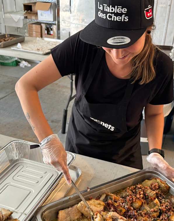 Dans une cuisine professionnelle, une personne vêtue d'un uniforme et d'une toque de chef noirs prépare des aliments sur un comptoir en acier inoxydable, avec divers équipements et ustensiles de cuisine visibles en arrière-plan.