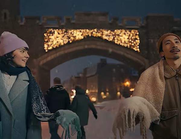 Deux personnes, un homme et une femme, se tiennent devant une arche ornée, illuminée par une lumière chaude, avec un paysage urbain visible en arrière-plan.