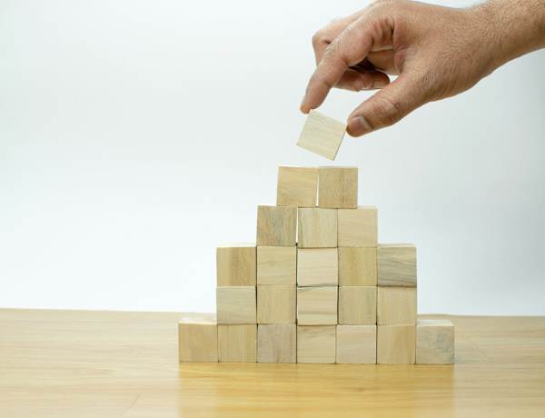 Une main dépose délicatement un bloc de bois au sommet d'une structure pyramidale composée d'autres blocs de bois, posée sur une table en bois.