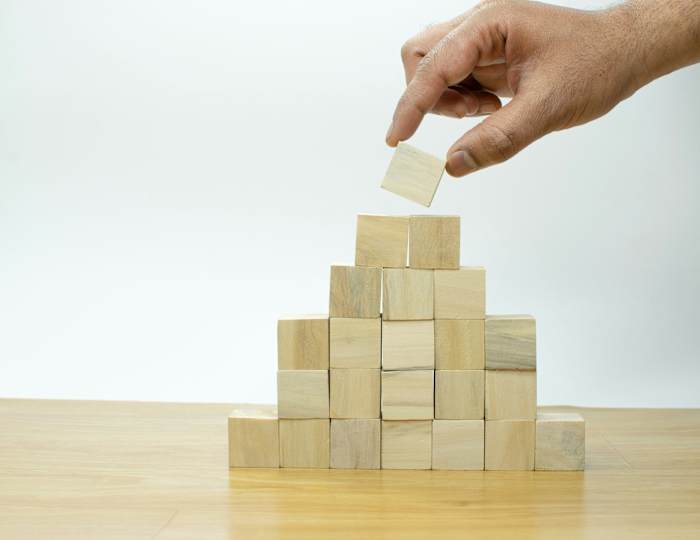 Une main dépose délicatement un bloc de bois au sommet d'une structure pyramidale composée d'autres blocs de bois, posée sur une table en bois.