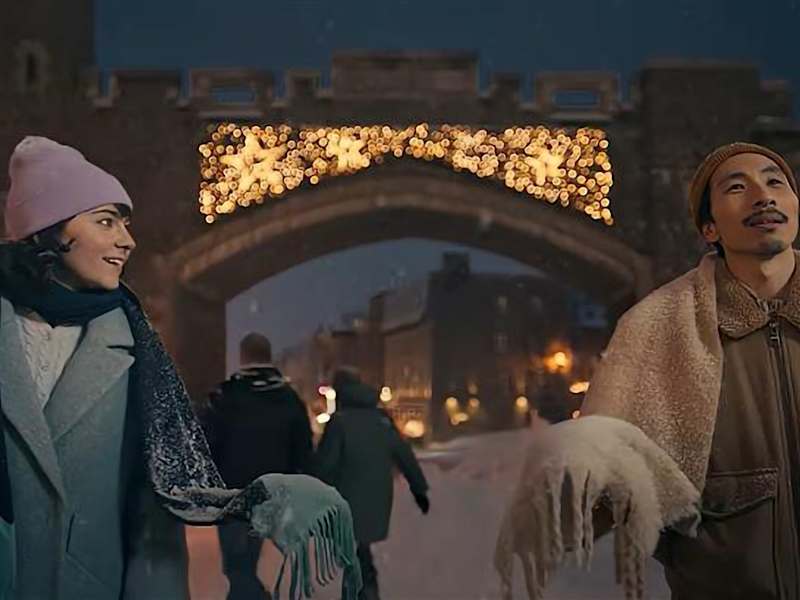 Deux personnes, un homme et une femme, se tiennent devant une arche ornée, illuminée par une lumière chaude, avec un paysage urbain visible en arrière-plan.