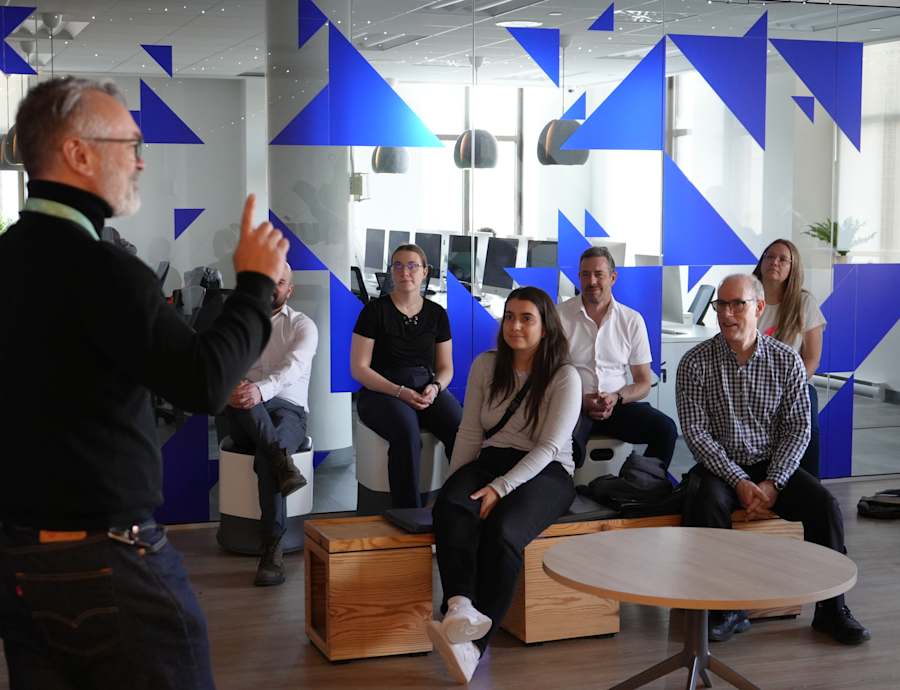Un homme en col roulé noir fait signe à un groupe de personnes assises dans un bureau moderne dont les murs sont ornés de motifs géométriques bleus.