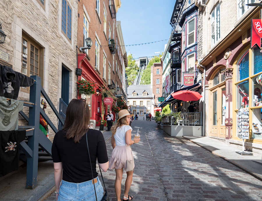Deux femmes marchent dans une ruelle pavée du Petit-Champlain en été, bordée de bâtiments en pierre et de commerces