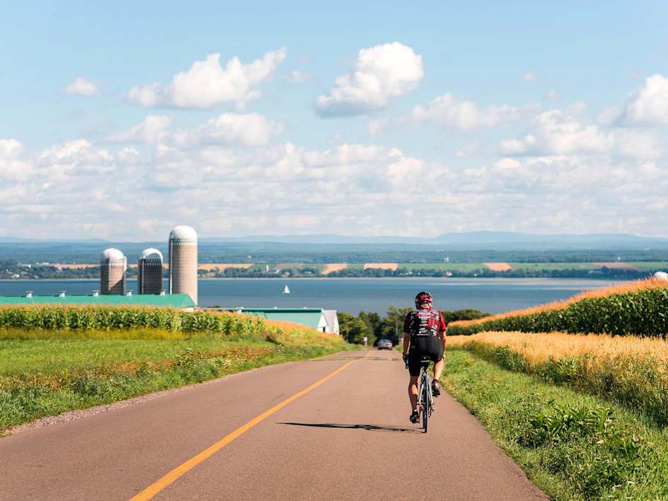 Un cycliste roule sur une piste pavée à travers un champ herbeux, avec un lac pittoresque et des bâtiments au loin visibles en arrière-plan.