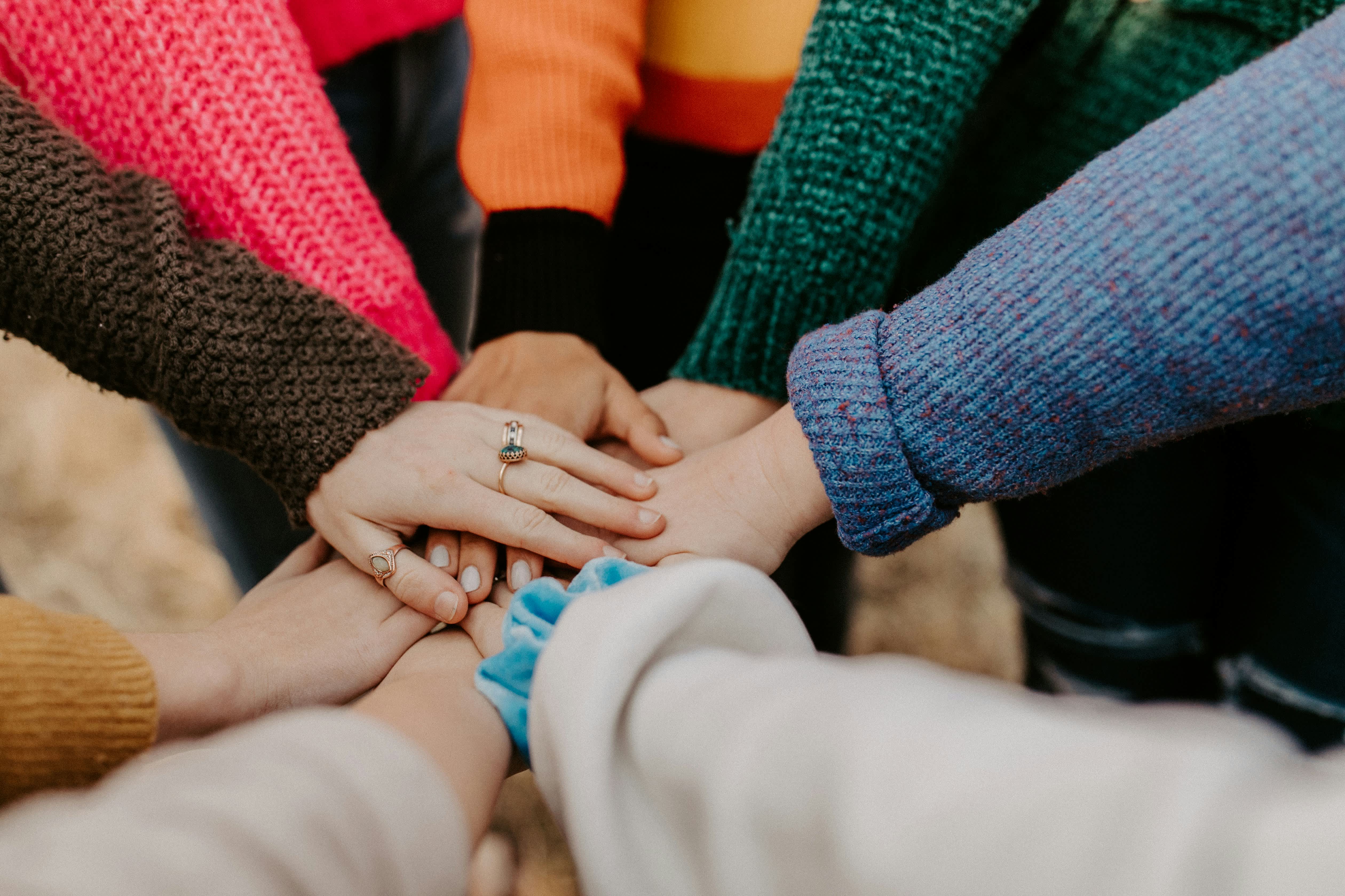 L'image montre plusieurs mains jointes, laissant apparaître des manches et des pulls colorés, suggérant un groupe de personnes réunies dans un esprit d'unité ou de soutien.